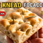 No knead rosemary garlic focaccia bread in a metal baking pan with crispy golden crust, fresh rosemary, and garlic topping